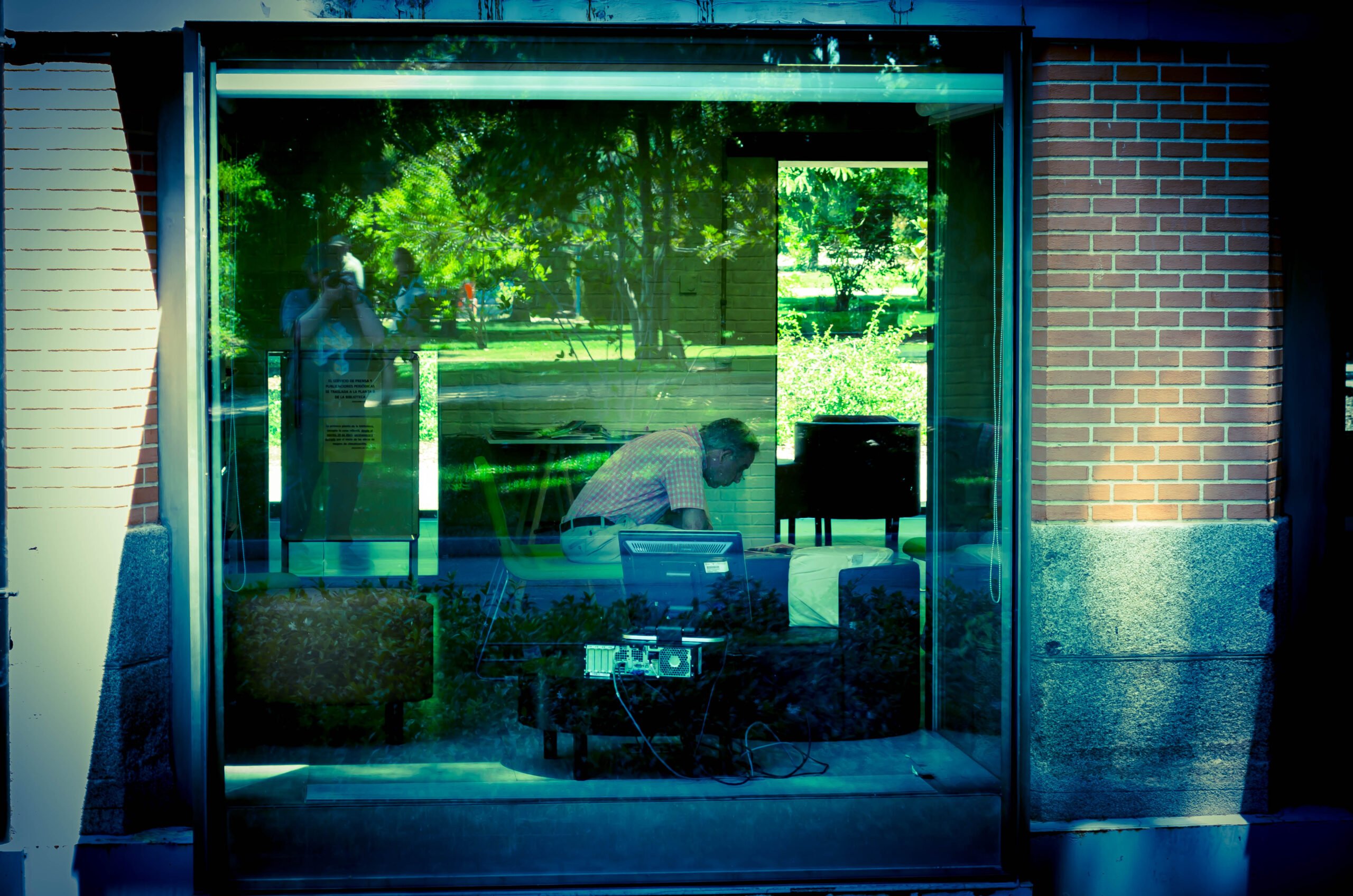 Man in house (picture taken from the street). Computer in foreground. Green reflection on window.