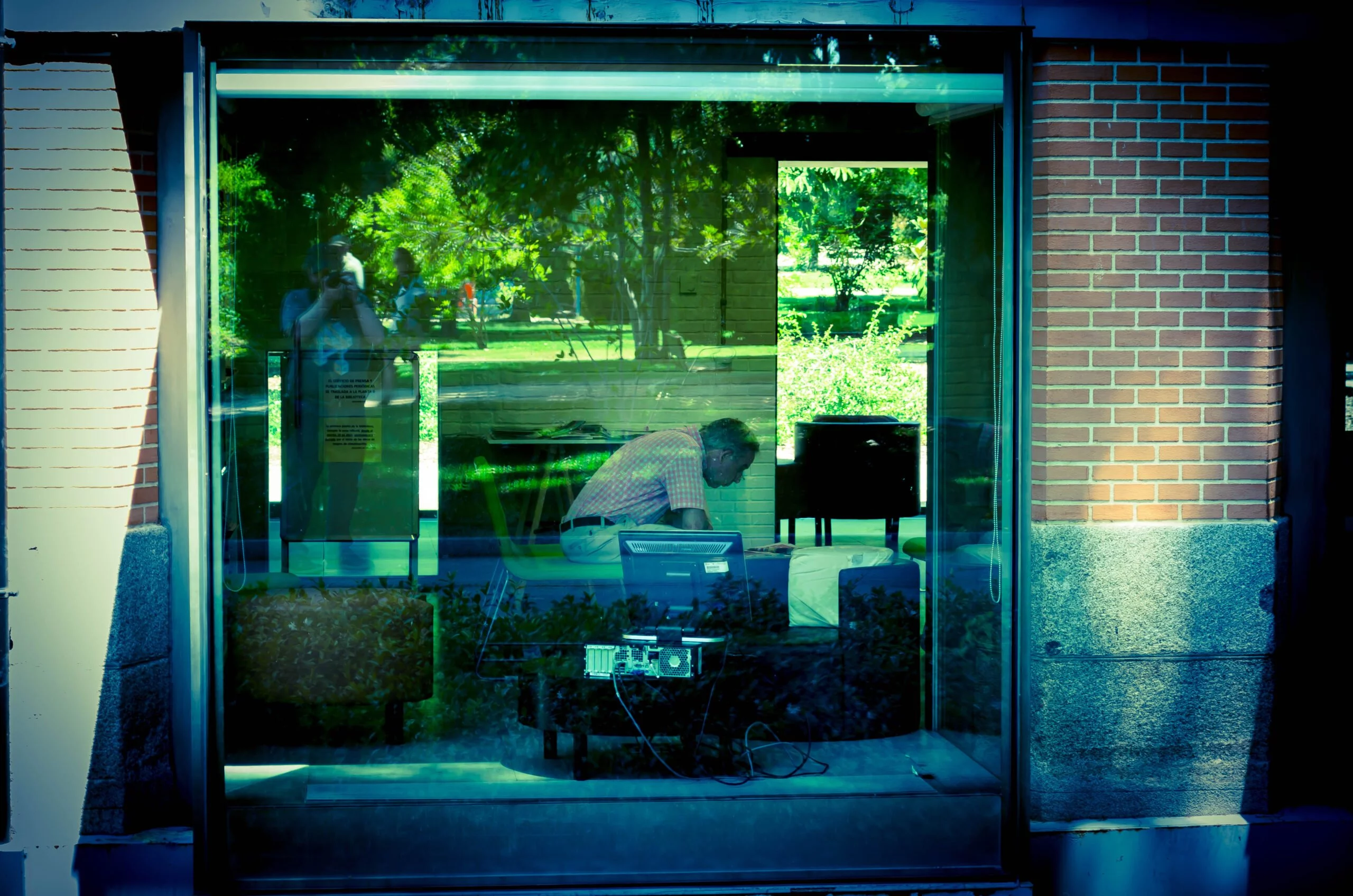 Man in house (picture taken from the street). Computer in foreground. Green reflection on window.